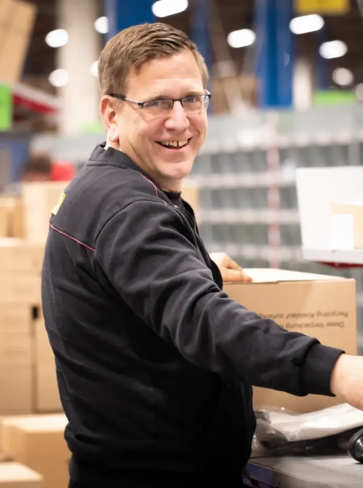 Ein Mann mit Brille und schwarzem Sweatshirt lächelt, während er in einem Lager oder einer Lagerhalle mit Kartons arbeitet.