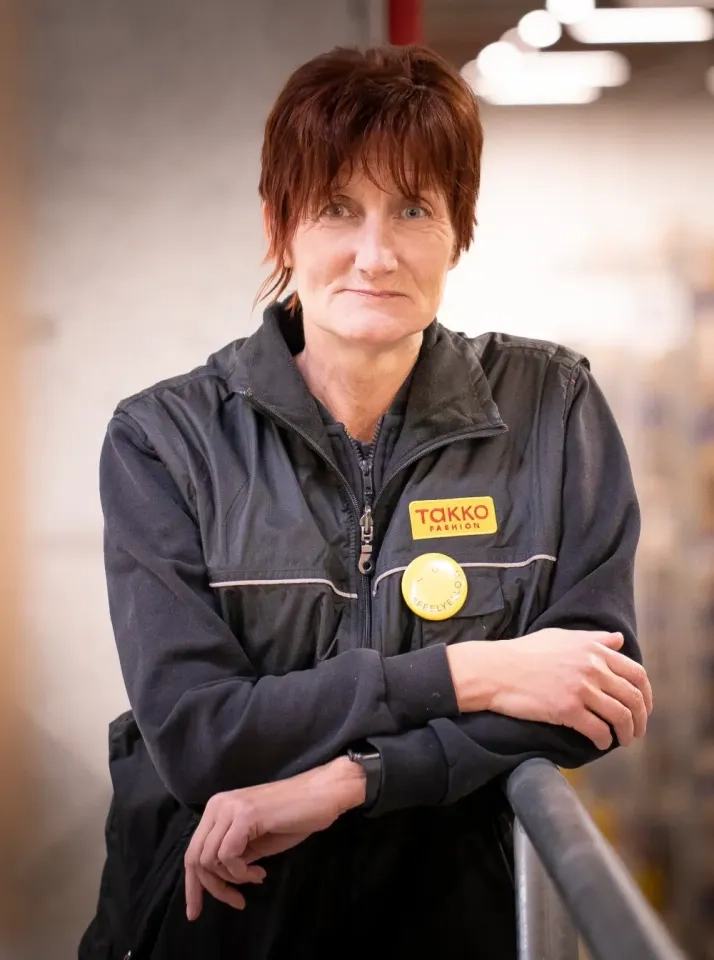 Eine Person mit kurzen braunen Haaren und einer schwarzen Arbeitsweste mit Takko Fashion-Logo steht im Innenbereich, lehnt an einem Geländer und blickt mit neutralem Gesichtsausdruck in die Kamera.