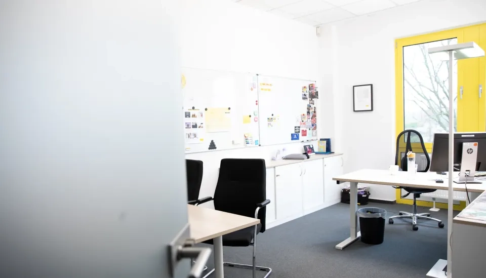Ein helles Büro mit weißen Wänden, einem gelb gerahmten Fenster, zwei Schreibtischen mit Computern, zwei schwarzen Stühlen, weißen Schränken, einem mit Notizen und Fotos bedeckten Whiteboard und verschiedenen Büromaterialien.