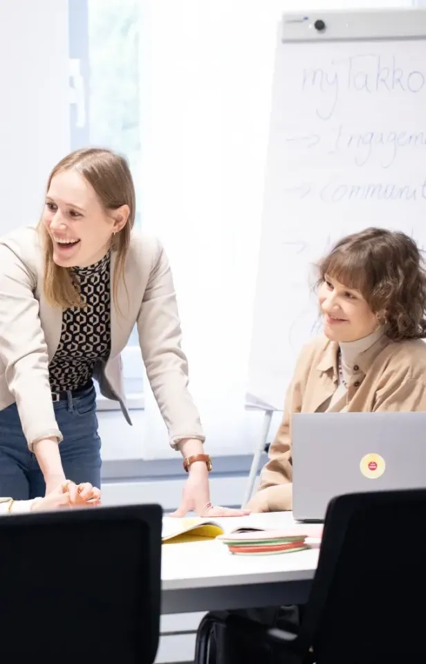 Zwei Frauen an einem Besprechungstisch; eine steht lächelnd und nach vorne gebeugt, die andere sitzt mit einem Laptop und lächelt ebenfalls. Auf dem Tisch liegen Papiere und Notizbücher, im Hintergrund ein Flipchart.