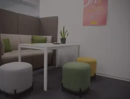 Ein moderner Loungebereich im Büro mit einem weißen Tisch, gepolsterten Sitzbänken mit grünen Kissen, drei runden Hockern in Grau, Grün und Gelb, einer Topfpflanze auf dem Tisch und einem rosa Poster an der Wand.