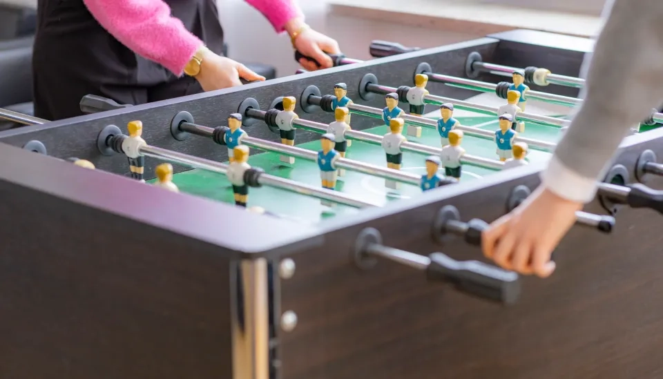 Zwei Personen spielen Tischfußball. Jeder Spieler steuert Griffe, die an Reihen von Miniaturfußballspielern auf einem grünen Feld in einem schwarzen Tischfußballspiel befestigt sind. Nur ihre Arme und Hände sind sichtbar.
