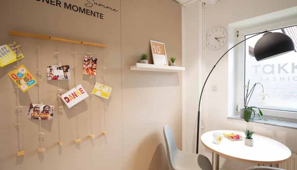 Eine gemütliche Ecke mit einem kleinen runden Tisch, zwei Stühlen, einer Wanduhr, einem Regal mit Pflanzen und einer Fotovitrine mit Postkarten und Fotos. Licht fällt durch ein Fenster mit einem TAKK FASHION-Aufkleber herein.