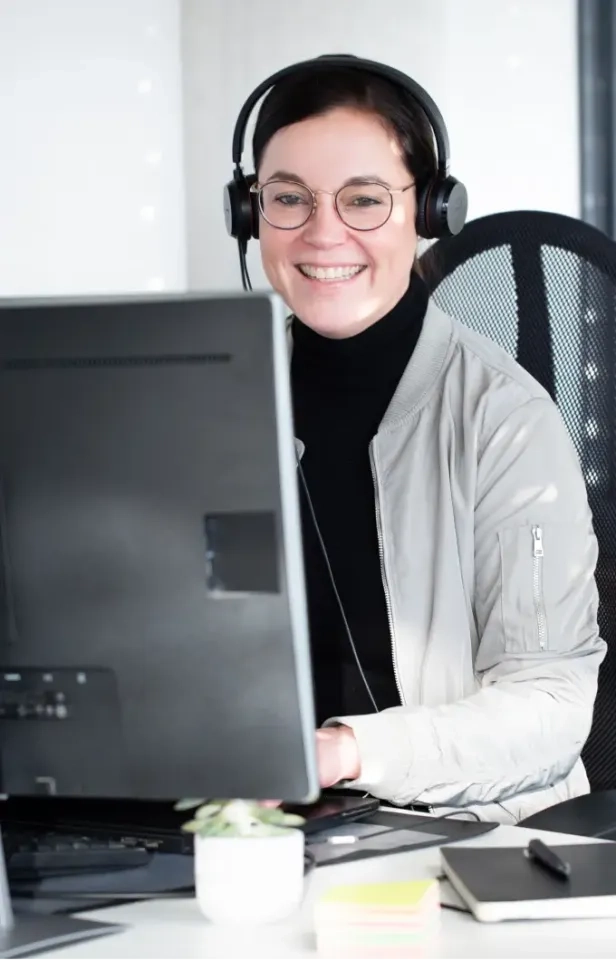 Eine lächelnde Person mit Brille und Kopfhörern sitzt an einem Schreibtisch und arbeitet am Computer. Vor ihr liegen eine kleine Pflanze, bunte Haftnotizen und ein Notizbuch auf dem Schreibtisch.