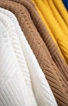 Nahaufnahme mehrerer in einer Reihe hängender Strickpullover in verschiedenen Farben und Texturen, darunter Weiß, Braun, Gelb und einem Hauch von Blau im Hintergrund.