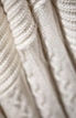 Nahaufnahme eines cremefarbenen Strickstoffs mit strukturierten Mustern und Zopfmustern. Das weiche Wollmaterial wirkt dick und gemütlich und hebt die komplizierten Details des Stricks hervor.