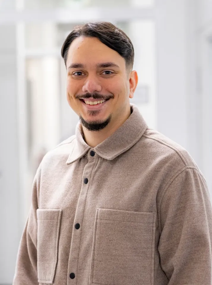 Ein Mann mit kurzen dunklen Haaren und Spitzbart lächelt in die Kamera. Er trägt eine beige Jacke mit Knöpfen und großen Vordertaschen und steht in einem hellen Innenraum mit unscharfem Hintergrund.