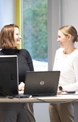 Zwei Frauen stehen und lächeln sich an einem Schreibtisch mit zwei Laptops in einem hellen Büro mit einem Fenster und Grün im Hintergrund.
