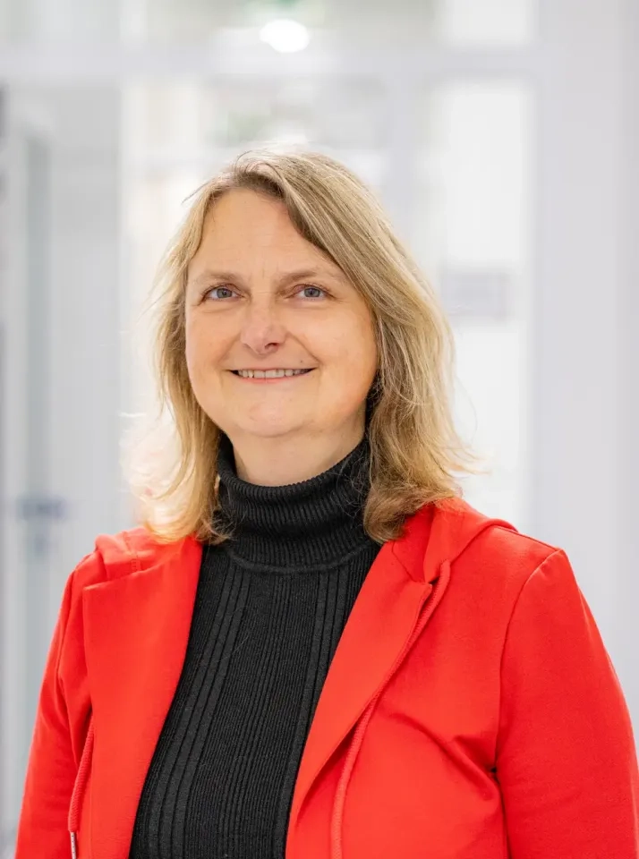 Eine Frau mit schulterlangem blondem Haar lächelt. Sie trägt einen schwarzen Rollkragenpullover und eine leuchtend rote Jacke und steht in einem Raum vor einem verschwommenen, lichtdurchfluteten Hintergrund.