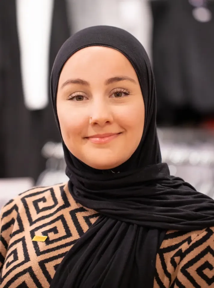 Eine Frau mit schwarzem Hijab und einem beige-schwarzen Oberteil mit geometrischem Muster lächelt in die Kamera. Sie trägt einen kleinen Nasenring und steht im Innenbereich. Im Hintergrund sind Kleiderständer unscharf zu sehen.