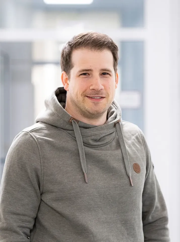 Ein Mann mit kurzen braunen Haaren und hellem Bartstoppeln lächelt sanft. Er trägt einen grauen Kapuzenpullover und steht in einem hellen, modernen Raum mit unscharfem Hintergrund.