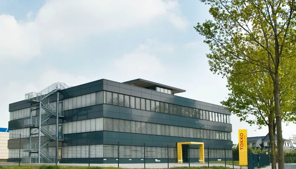 Ein modernes, dreistöckiges Bürogebäude mit dunklen Glaspaneelen und einem gelb umrahmten Eingang. Im Vordergrund sind unter einem leicht bewölkten Himmel ein Baum und ein gelbes Schild mit der Aufschrift „TOKKO“ zu sehen.