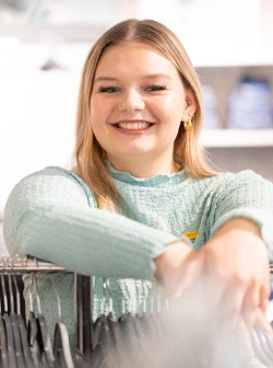 Eine junge Frau mit langen blonden Haaren und einem hellblauen strukturierten Oberteil lächelt, während sie in einem hell erleuchteten Geschäft an einer Kleiderstange lehnt.
