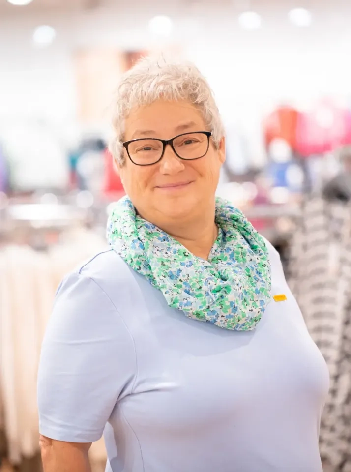 Eine ältere Frau mit kurzen grauen Haaren und Brille lächelt in die Kamera. Sie trägt ein hellblaues Oberteil und einen blau-grünen Blumenschal und steht in einem hell erleuchteten Bekleidungsgeschäft.