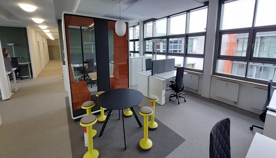 Moderner Büroraum mit großen Fenstern, einem runden schwarzen Tisch, umgeben von gelben Hockern, einer halbprivaten Besprechungskabine mit orangefarbenen Sitzen, Schreibtischen mit Computern und einer arbeitenden Person im Hintergrund.