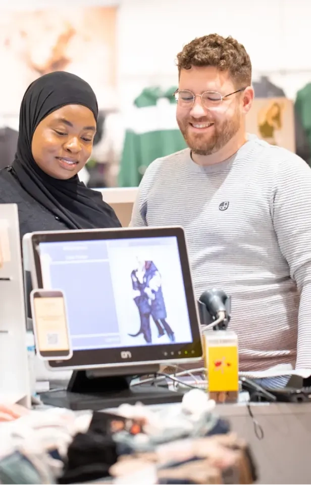 Zwei Einzelhandelsmitarbeiterinnen, eine mit Hijab, die andere mit Brille und Bart, lächeln, während sie an der Kasse auf einen Computerbildschirm schauen. Im Hintergrund sind Kleidung und Regale zu sehen.