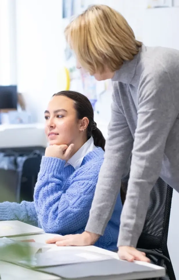 Eine Frau in einem blauen Pullover sitzt an einem Schreibtisch, stützt ihr Kinn auf ihre Hand und sieht nachdenklich aus, während eine andere Person neben ihr steht, sich über den Schreibtisch beugt und mit ihr spricht.