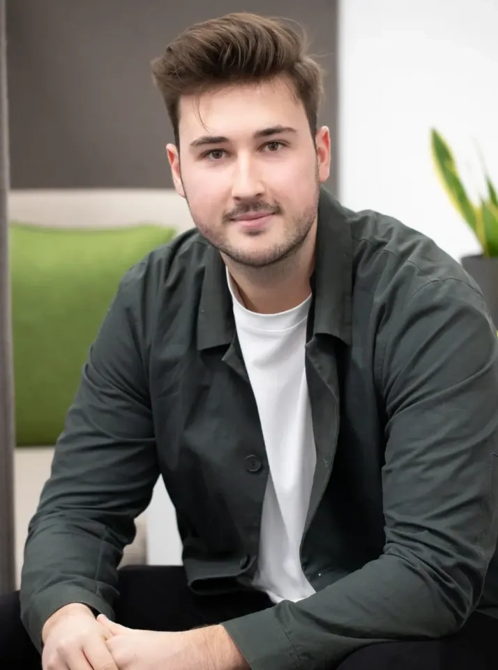 Ein junger Mann mit kurzen braunen Haaren und hellem Bartstoppeln sitzt drinnen. Er trägt eine dunkelgrüne Jacke über einem weißen T-Shirt und blickt mit einem leichten Lächeln in die Kamera. Im Hintergrund liegen ein grünes Kissen und eine Pflanze.