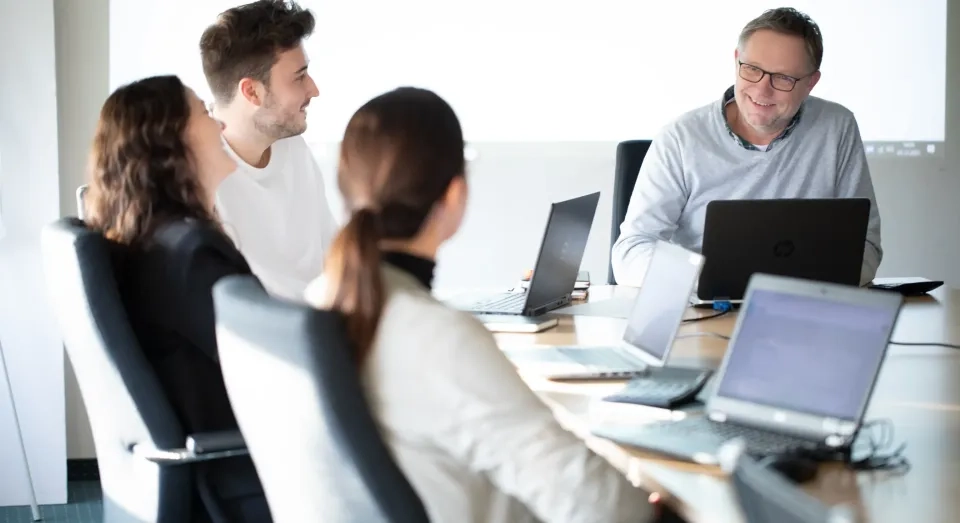 Vier Personen sitzen mit Laptops um einen Konferenztisch und sind in einer Besprechung. Ein lächelnder Mann scheint die Diskussion zu leiten. Die Atmosphäre wirkt kooperativ und professionell.