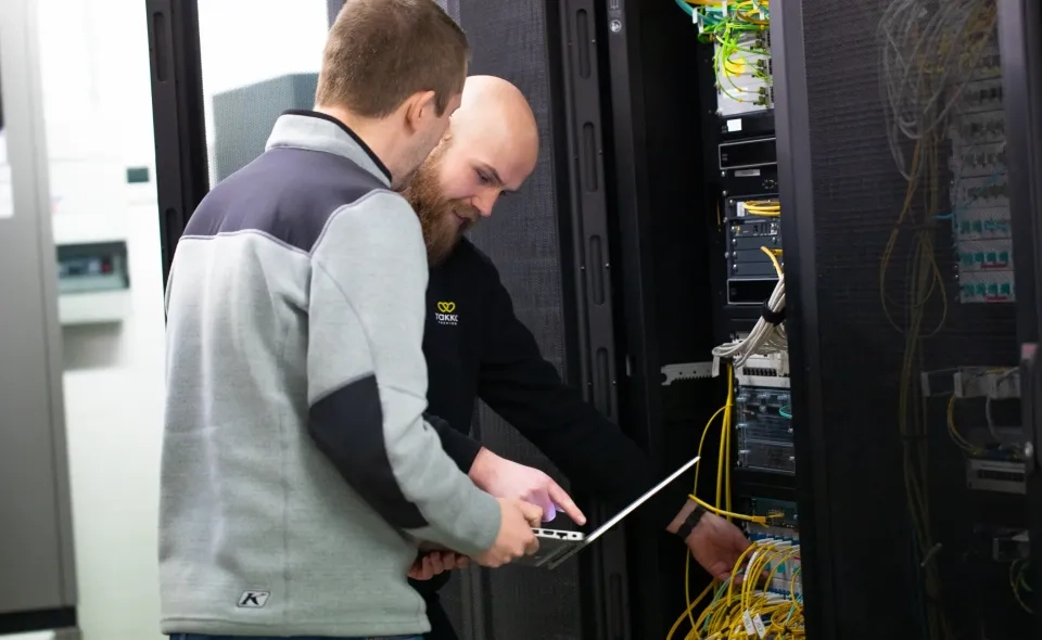 Zwei Männer arbeiten in einem Serverraum zusammen. Einer hält einen Laptop, während der andere die Kabel verwaltet, die mit einem Server-Rack voller Netzwerkgeräte verbunden sind.