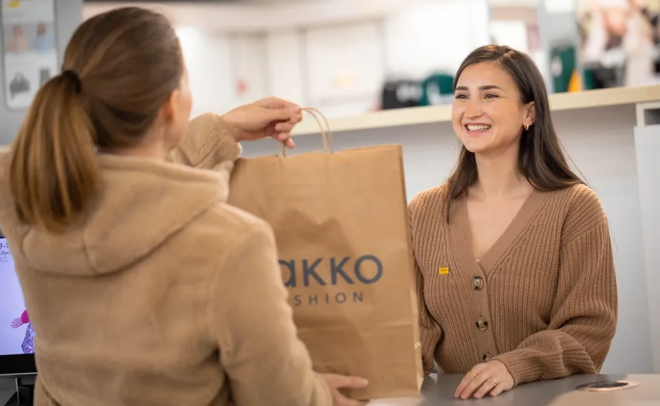 Eine lächelnde Frau an der Kasse erhält von einer anderen Person eine Papiereinkaufstüte. Beide tragen hellbraune Pullover. Auf der Tüte steht der Aufdruck „AKKO FASHION“. Die Szene scheint in einem Bekleidungsgeschäft zu sein.
