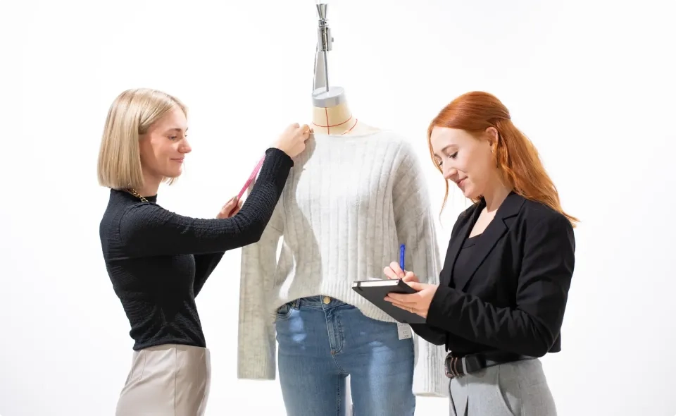 Zwei Frauen arbeiten gemeinsam an einem Modeprojekt; die eine misst mit einem Maßband einen Pullover auf einer Schneiderpuppe, während die andere konzentriert und lächelnd Notizen in einem Notizblock macht.