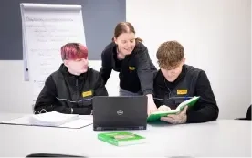 Ein Lehrer steht da und zeigt auf ein Lehrbuch, während er zwei Schülern hilft, die in einem Klassenzimmer an einem Tisch mit offenen Notizbüchern und einem Laptop sitzen.