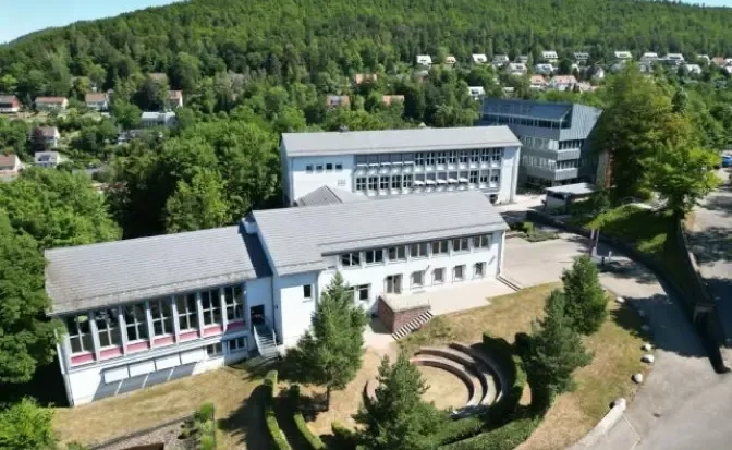 Luftaufnahme eines modernen Schulcampus mit weißen Gebäuden, einem Freiluft-Amphitheater und umgebenden Bäumen. Im Hintergrund sind Wohnhäuser und ein bewaldeter Hang zu sehen.