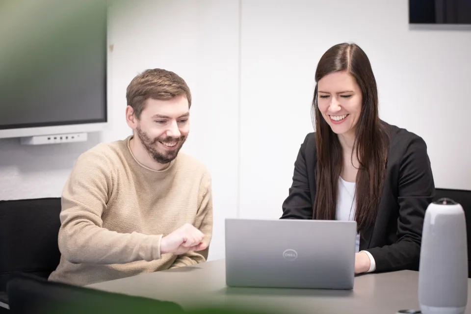 Zwei Personen sitzen lächelnd an einem Tisch und schauen auf einen Laptop-Bildschirm. Eine Person zeigt auf den Bildschirm, während die andere tippt. Im Hintergrund ist ein Monitor an der Wand montiert.