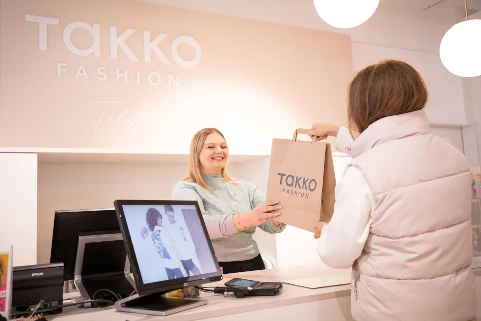 Eine lächelnde Kassiererin reicht einem Kunden in einem TAKKO Fashion Store eine Einkaufstasche. Das Logo des Geschäfts ist an der Wand und auf der Tasche zu sehen. Auf der Theke stehen ein Computer und ein Bezahlterminal.