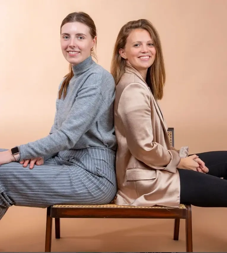Zwei Frauen sitzen Rücken an Rücken auf einer Bank vor beigem Hintergrund. Die eine trägt einen grauen Pullover und gestreifte Hosen, die andere einen hellbraunen Blazer und schwarze Hosen. Beide lächeln und blicken in die Kamera.