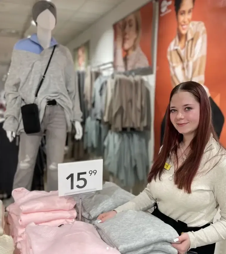 Eine junge Frau mit langen braunen Haaren steht in einem Bekleidungsgeschäft neben gefalteten Pullovern und einem Preisschild mit der Aufschrift „15,99“. Im Hintergrund ist eine Schaufensterpuppe in grauem Outfit zu sehen. Das Geschäft präsentiert Kleidung in neutralen Farben.