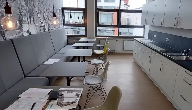 Moderner Pausenraum im Büro mit grau gepolsterter Bank, kleinen Tischen, nicht zueinander passenden Stühlen, hängenden Glühbirnen, Küchenbereich mit Schränken und Spüle, Fenstern mit Blick auf die Stadt und Papieren auf einem Tisch.