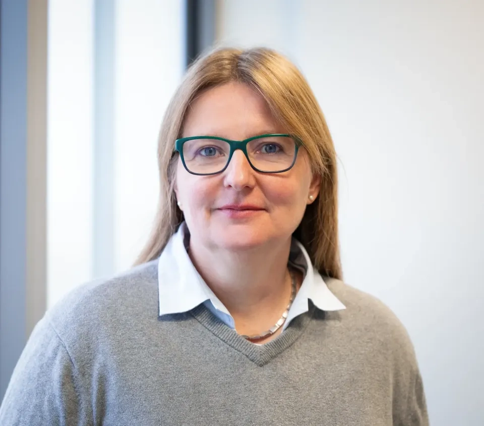 Eine Frau mit glattem blondem Haar, einer blaugrünen Brille, einem weißen Hemd mit Kragen und einem hellgrauen Pullover steht vor einem sanft beleuchteten Hintergrund mit großen Fenstern.