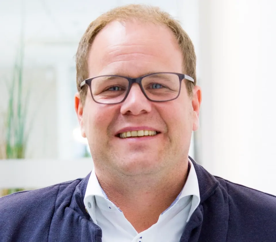 Ein Mann mit kurzen hellbraunen Haaren und Brille lächelt in die Kamera. Er trägt ein weißes Hemd mit Kragen und eine dunkle Jacke. Der Hintergrund ist hell und leicht unscharf, was auf eine Innenaufnahme schließen lässt.