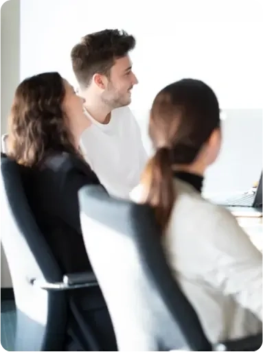 Drei Personen sitzen auf Bürostühlen an einem Konferenztisch, blicken in die gleiche Richtung und scheinen in eine Besprechung oder Diskussion vertieft zu sein. Die Szene ist hell mit natürlichem Licht beleuchtet.