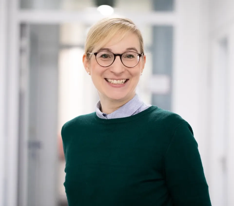 Eine Person mit kurzen blonden Haaren und Brille, die einen grünen Pullover über einem Hemd mit Kragen trägt, lächelt, während sie in einem hell erleuchteten Flur mit weißen Wänden und offenen Türen steht.