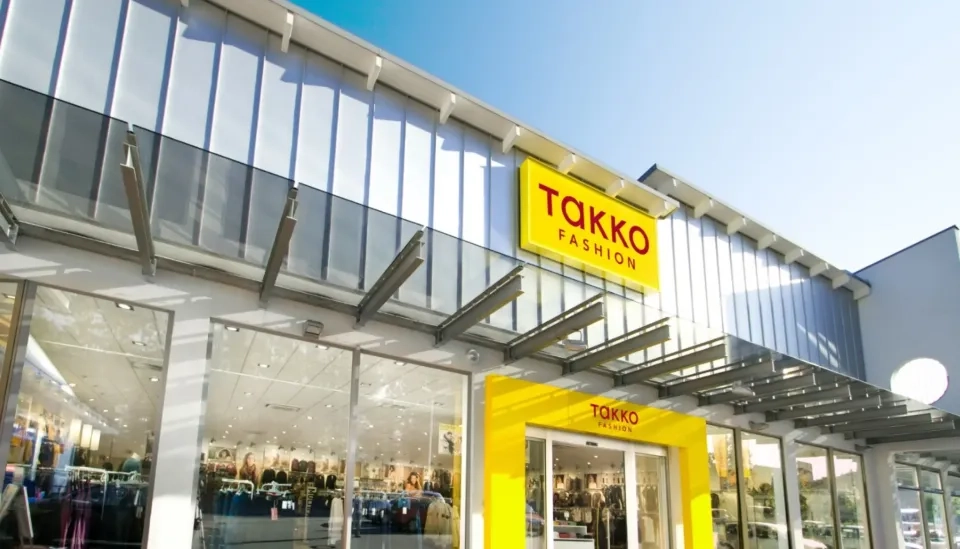 Eine moderne Ladenfront mit großen Glasfenstern und einem gelben Takko Fashion-Schild über dem Eingang, in dem sich der strahlend blaue Himmel spiegelt. Im hellen Laden sind die Kleiderauslagen gut sichtbar.