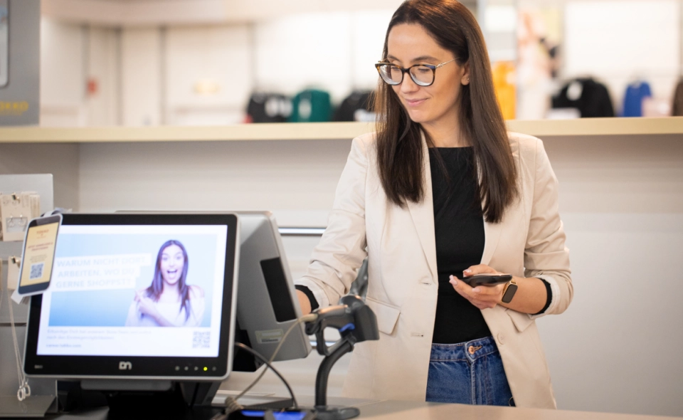 Eine Frau mit langen braunen Haaren und Brille bedient einen Touchscreen an einer Kasse und hält dabei ihr Smartphone in der Hand. Auf dem Monitor neben ihr sind eine lächelnde Frau und ein Text zu sehen. Im Hintergrund ist ein helles Einzelhandelsgeschäft zu sehen.