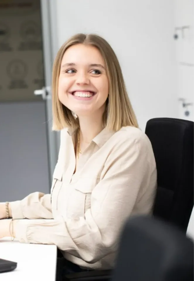 Eine junge Frau mit glattem blondem Haar sitzt an einem Schreibtisch, lächelt strahlend und blickt nach links. Sie trägt eine hellbeige Bluse und scheint sich in einer modernen Büroumgebung zu befinden.