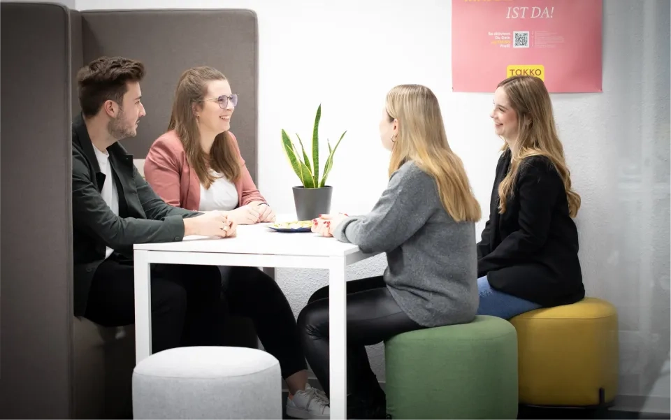 Vier Personen sitzen in einem modernen Büro um einen weißen Tisch, unterhalten sich und lächeln. Eine Topfpflanze steht auf dem Tisch, bunte Hocker dienen als Sitzgelegenheiten. Im Hintergrund hängt ein rosa Poster an der Wand.