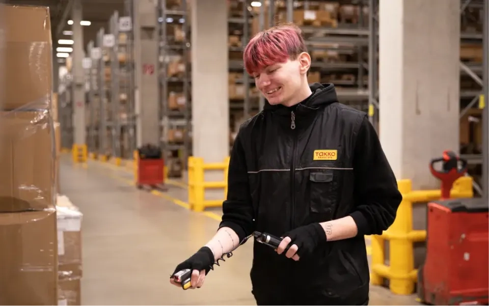Eine Person mit kurzen rotbraunen Haaren lächelt, während sie in einem Lager arbeitet. Sie hält einen Scanner in der Hand und trägt Handschuhe und eine schwarze Weste mit einem TAKKO-Logo. Im Hintergrund sind Regale und Kisten zu sehen.