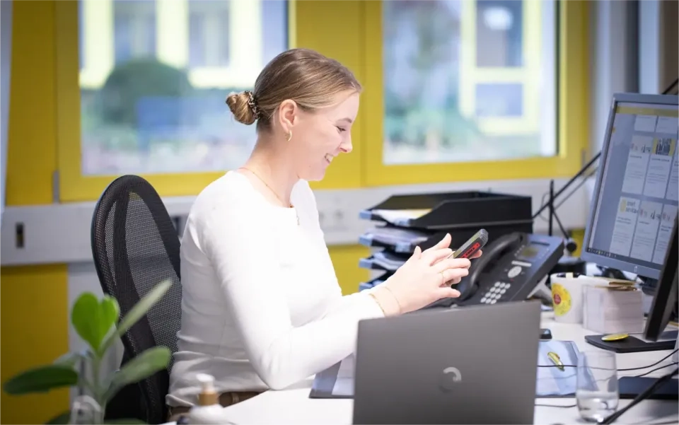Eine Frau sitzt lächelnd an einem Schreibtisch und benutzt ihr Smartphone. Sie ist umgeben von Bürogeräten wie Laptop, Telefon, Papieren und einer Pflanze. Im Hintergrund sind leuchtend gelbe Fensterrahmen zu sehen.