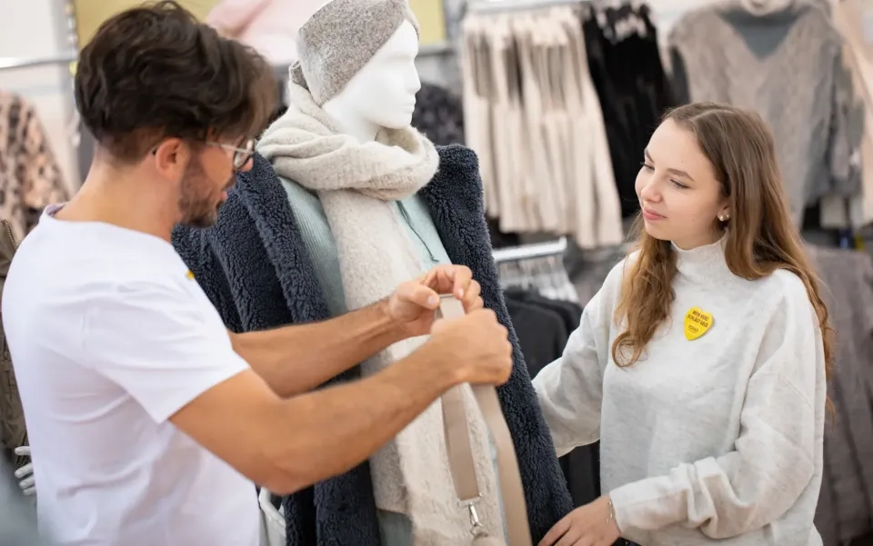Ein Mann und eine Frau richten in einem Bekleidungsgeschäft eine Handtasche an einer Schaufensterpuppe in Winterkleidung. Die Schaufensterpuppe trägt eine Strickmütze, einen Schal und einen Mantel. Die Frau und der Mann scheinen zusammenzuarbeiten.