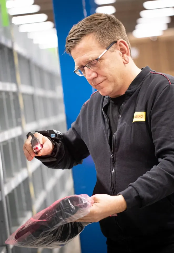 Ein Mann mit Brille scannt in einem Lagerhaus neben Metallregalen einen verpackten Artikel mit einem tragbaren Barcode-Scanner.