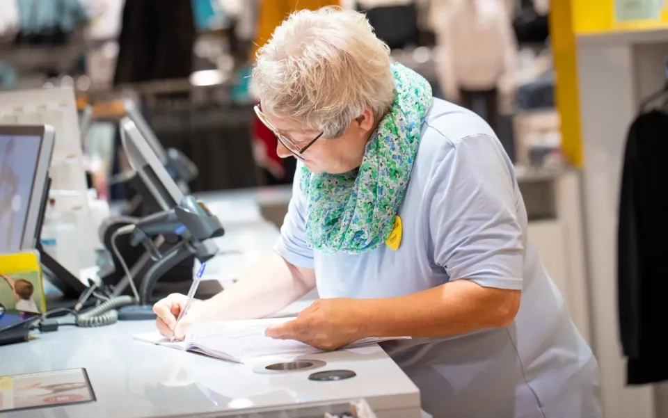 Eine ältere Frau mit kurzen grauen Haaren und Brille schreibt in ein Notizbuch an einer Ladentheke. Sie trägt ein hellblaues Hemd und einen grün gemusterten Schal und hat einen Computer und ein Telefon in der Nähe.