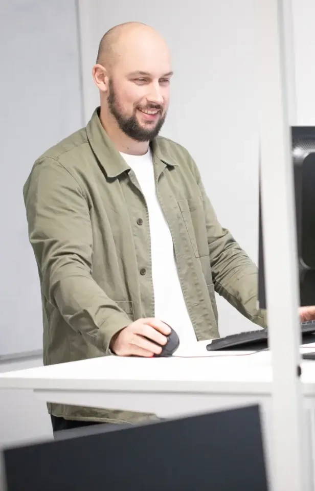 Ein Mann mit Bart und rasiertem Kopf, der eine grüne Jacke und ein weißes Hemd trägt, steht an einem Schreibtisch, bedient eine Computermaus und lächelt auf einen Monitor in einem hellen, modernen Büro.
