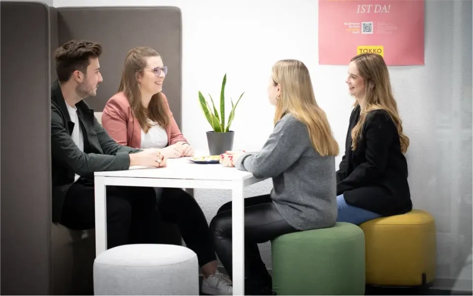 Vier Personen sitzen um einen weißen Tisch, unterhalten sich und lächeln. Eine Topfpflanze steht auf dem Tisch. Sie sitzen auf runden Hockern in einem hellen, modernen Büro. Im Hintergrund hängt ein rosa Poster an der Wand.