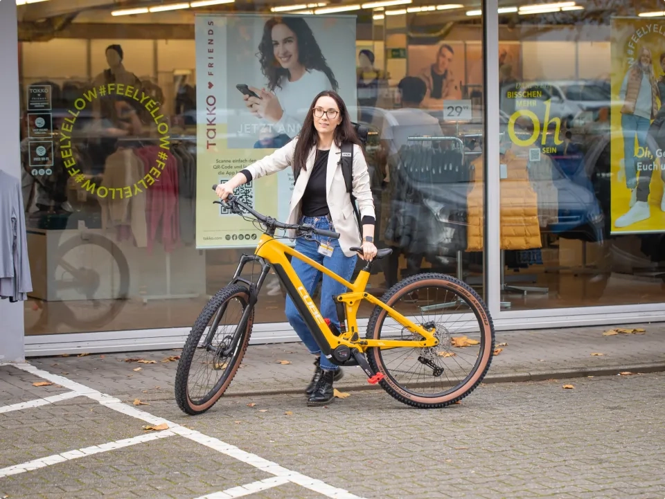 Eine Frau mit Brille und leichter Jacke steht vor einem Geschäft und hält ein gelb-blaues Fahrrad in der Hand. In den Schaufenstern sind Plakate und Schaufensterpuppen zu sehen, in den Scheiben spiegeln sich geparkte Autos.