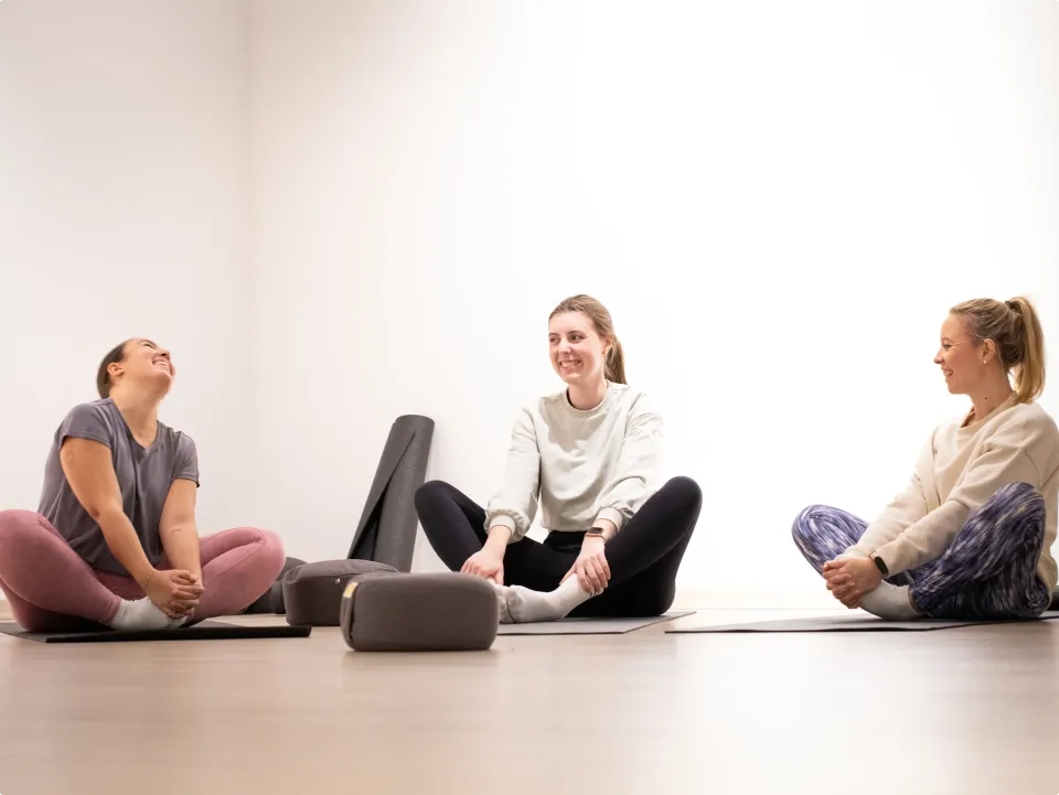 Drei Frauen sitzen in einem hellen Raum auf Yogamatten und lächeln und lachen gemeinsam. Yoga-Utensilien wie Bolster und Matten liegen um sie herum auf dem Boden. Sie wirken entspannt und tragen bequeme Trainingskleidung.